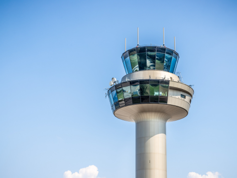 A modern airport control tower with large blue-tinted windows stands against a clear blue sky, representing advanced Kontrollmanagement. A few small clouds linger near the bottom of the image.