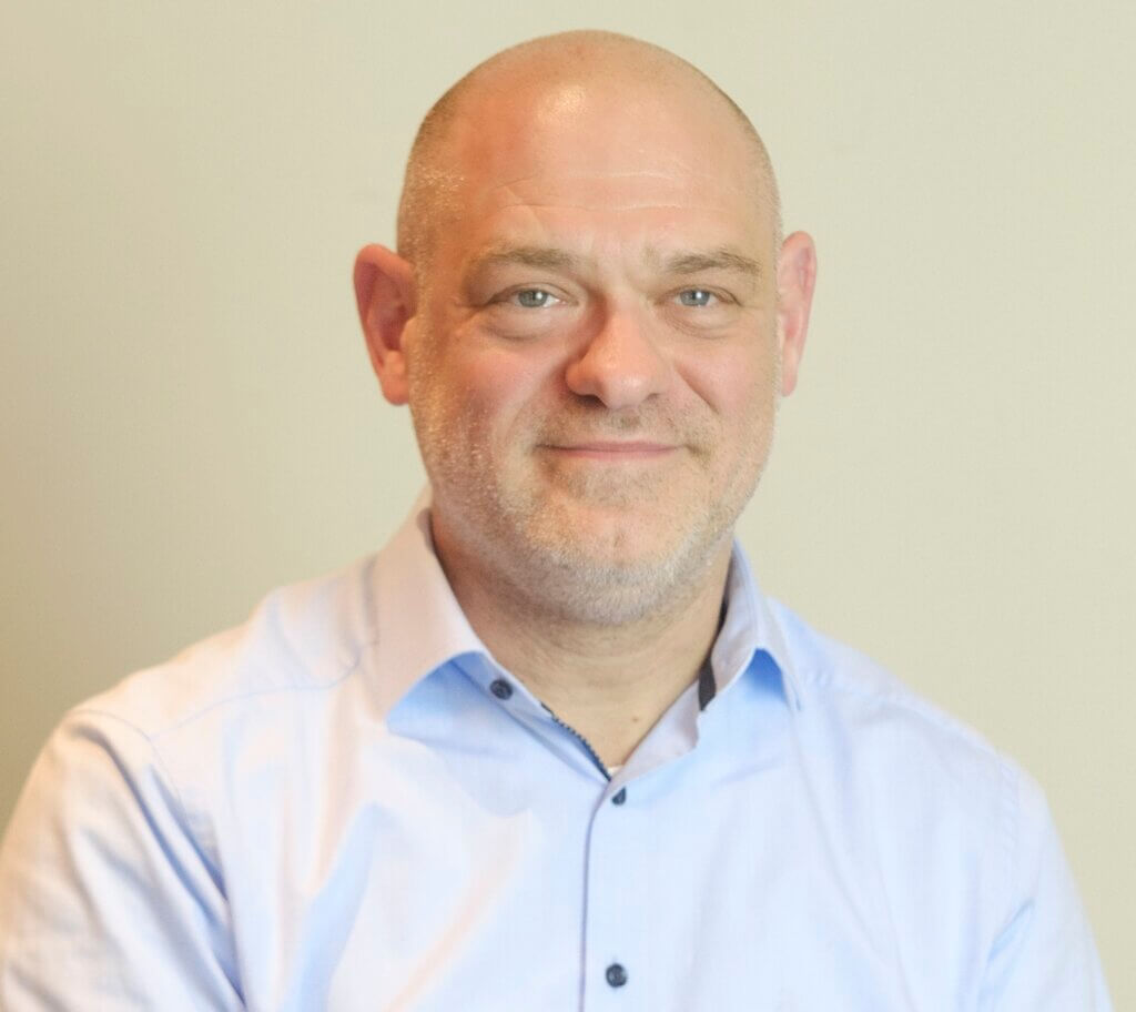 Ein Mann mittleren Alters mit Glatze und leichten Bartstoppeln lächelt sanft. Er trägt ein hellblaues Firmen-Button-up-Hemd und steht vor einem schlichten, hellen Hintergrund.