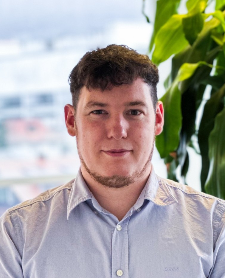 Ein Mann mit kurzem dunklem Haar und gestutztem Bart, der ein hellblaues Hemd mit Kragen trägt, steht in einem Haus vor einer großen Blattpflanze und einem hellen, unscharfen Firmenhintergrund.