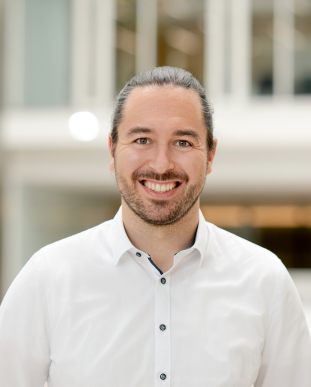 Ein Mann mit langen, zurückgebundenen braunen Haaren, Bart und Schnurrbart, der ein weißes Button-up-Hemd trägt, lächelt, während er in einem Haus vor einem unscharfen Firmenhintergrund steht.